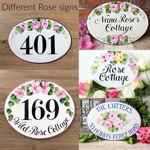 May include: Four oval-shaped ceramic signs with hand-painted rose designs. The signs feature house numbers, names, and addresses. The signs are white with pink, yellow, and green floral accents.