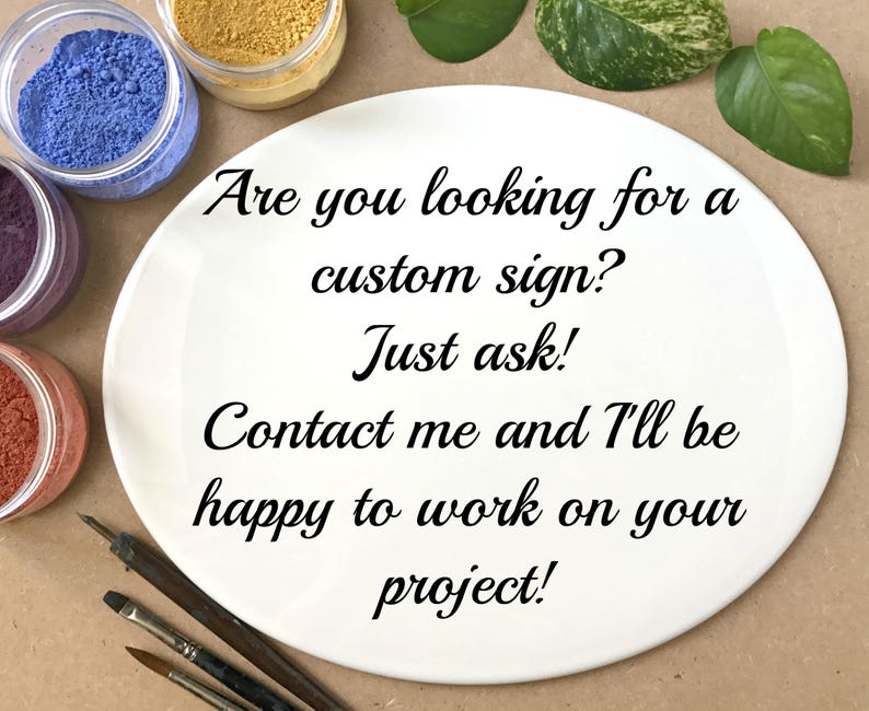 May include: A white plate with the text "Are you looking for a custom sign? Just ask! Contact me and I'll be happy to work on your project!" written in black cursive font. The plate is surrounded by small jars of colorful powder and paintbrushes.