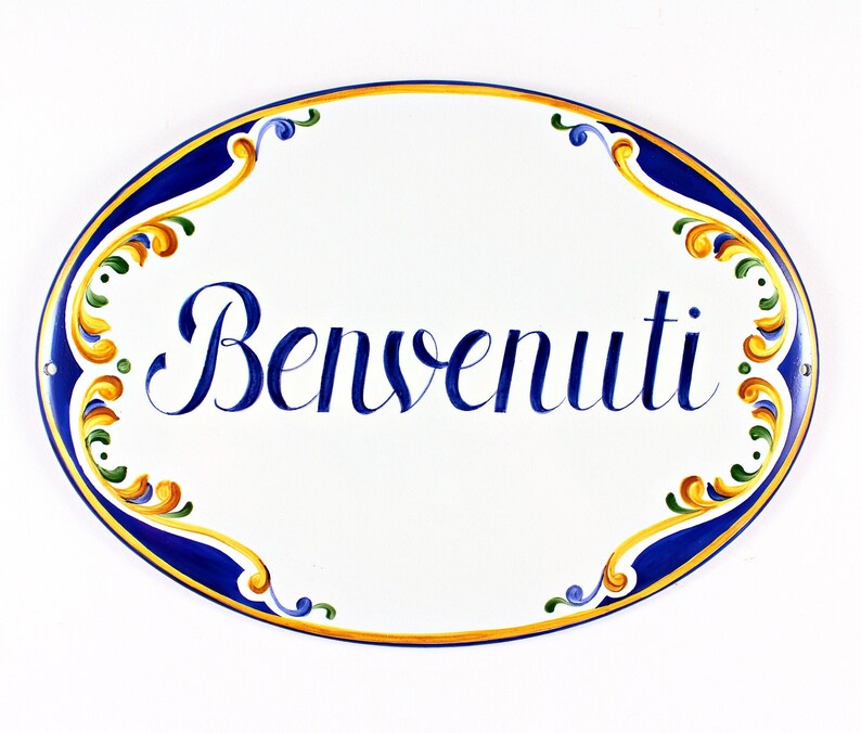 Oval ceramic sign with the word "Benvenuti" in elegant blue script. The sign has a white background with a blue and gold hand-painted border. The border features ornate floral accents.