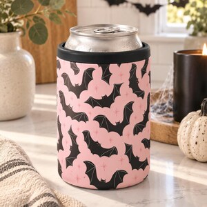 May include: A cylindrical metal can cooler featuring a repeating pattern of black bats on a light pink background, accented with small pink starburst shapes. The cooler has a solid black rim at the top and holds a standard silver aluminum beverage can.