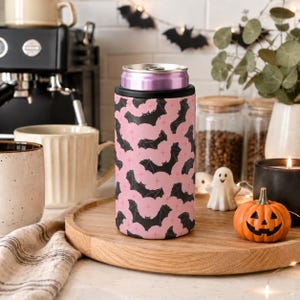 May include: A cylindrical can cooler featuring a repeating pattern of black bats and small pink stars on a light pink background. The cooler has a black rim at the top and holds a metallic purple beverage can, standing upright on a round wooden tray.