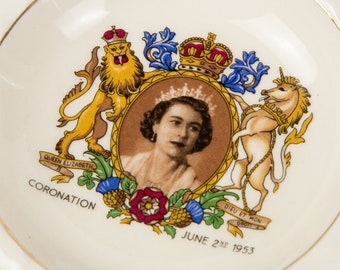 Puede incluir: Un plato de cerámica blanco con un borde dorado presenta una imagen colorida de la coronación de la reina Isabel II. La imagen incluye un león, un unicornio y una corona. El texto "Queen Elizabeth II Coronation June 2nd 1953" está impreso en el plato.