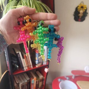Puede incluir: Cuatro figuritas de lagarto de cuentas de colores, cada una de un color diferente del arco&iacute;ris: naranja, verde, azul y morado. Los lagartos est&aacute;n hechos de peque&ntilde;as cuentas redondas y tienen un acabado brillante e iridiscente.