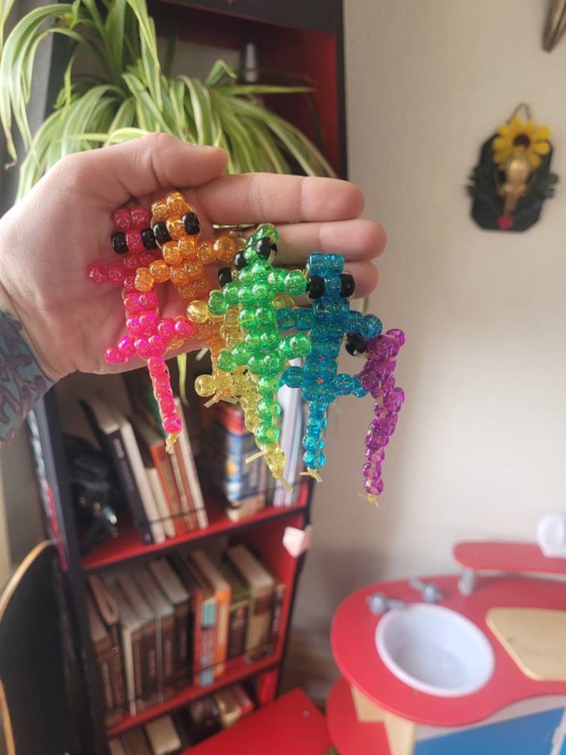 Puede incluir: Cuatro figuritas de lagarto de cuentas de colores, cada una de un color diferente del arco&iacute;ris: naranja, verde, azul y morado. Los lagartos est&aacute;n hechos de peque&ntilde;as cuentas redondas y tienen un acabado brillante e iridiscente.
