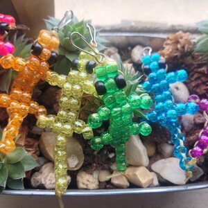 Puede incluir: Cinco figuritas de lagarto de cuentas en un arco&iacute;ris de colores, incluyendo rosa, naranja, amarillo, verde y azul. Los lagartos est&aacute;n hechos de peque&ntilde;as cuentas de colores y est&aacute;n enhebrados en un alambre.