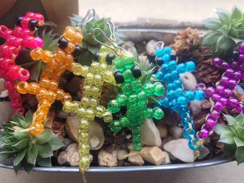 Puede incluir: Cinco figuritas de lagarto de cuentas en un arco&iacute;ris de colores, incluyendo rosa, naranja, amarillo, verde y azul. Los lagartos est&aacute;n hechos de peque&ntilde;as cuentas de colores y est&aacute;n enhebrados en un alambre.