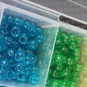 Puede incluir: Un contenedor de almacenamiento de pl&aacute;stico transparente con m&uacute;ltiples compartimentos llenos de cuentas coloridas y brillantes. Las cuentas est&aacute;n dispuestas en filas por color, con cuentas moradas, azules, verdes, amarillas, naranjas y rojas.