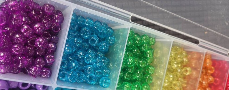 Puede incluir: Un contenedor de almacenamiento de pl&aacute;stico transparente con m&uacute;ltiples compartimentos llenos de cuentas coloridas y brillantes. Las cuentas est&aacute;n dispuestas en filas por color, con cuentas moradas, azules, verdes, amarillas, naranjas y rojas.