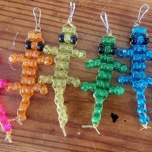 Puede incluir: Seis llaveros de lagarto de cuentas de colores, cada uno con ojos negros. Los lagartos son rosa, naranja, amarillo, verde, azul y morado.