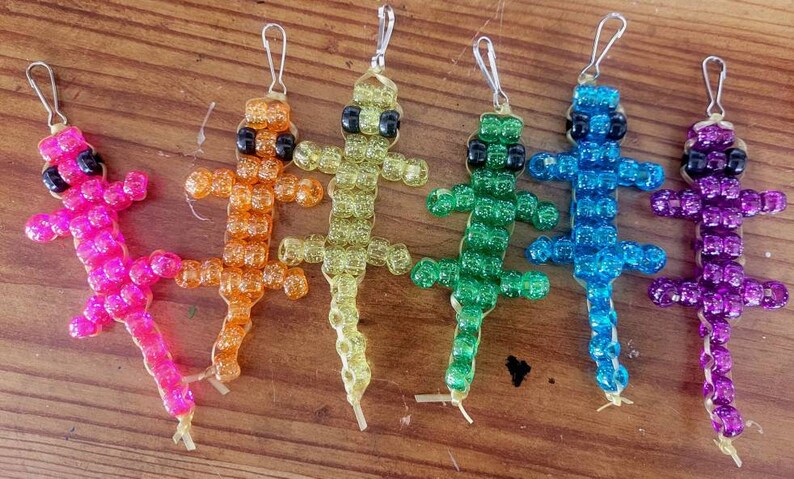 Puede incluir: Seis llaveros de lagarto de cuentas de colores, cada uno con ojos negros. Los lagartos son rosa, naranja, amarillo, verde, azul y morado.