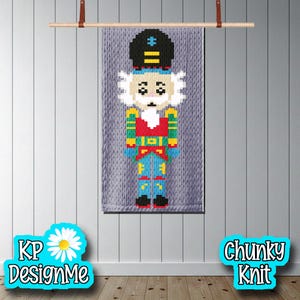 Könnte beinhalten: Ein farbenfrohes, pixeliges Nussknacker-Wandbild. Das Design zeigt einen schwarzen Hut, weißes Haar und eine rote, grüne und blaue Uniform. Das Kunstwerk wird auf einem grauen, grob gestrickten Hintergrund präsentiert und ist an einer Holzstange aufgehängt.