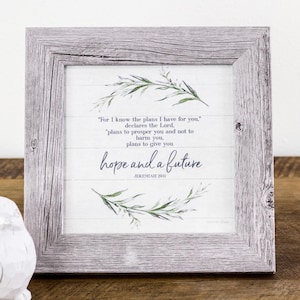 May include: A square wall art piece in a weathered gray wood-look frame. The white background features horizontal lines and green watercolor leaf illustrations. The text reads: For I know the plans I have for you, declares the Lord, hope and a future.