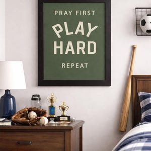 Black color frame of a Christian sports framed wall art with the phrase Pray First Play Hard Repeat on a green background in a vertical layout. Pictured in a boys room.