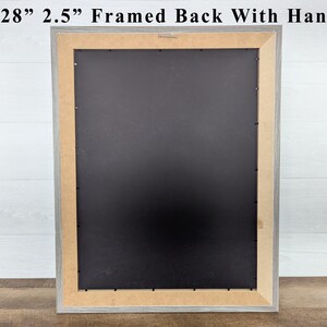May include: A framed artwork back with a hanger. The frame is 22x28 inches and 2.5 inches wide. The frame is gray with a black backing and a light brown interior border. The text at the top reads: 22x28" 2.5" Framed Back With Hanger.