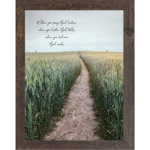 May include: A framed wall art piece featuring a dirt path winding through a green wheat field under a cloudy sky. The text reads: When you pray God listens, when you listen God talks, when you believe God works. The frame is dark brown wood.