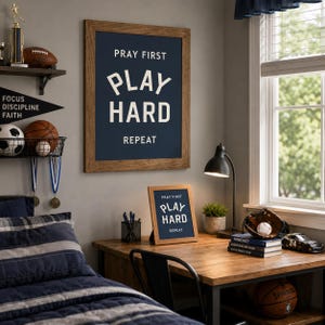 May include: A wall-mounted framed print and a matching desk-top framed print, both featuring a dark blue background with white text that reads PRAY FIRST, PLAY HARD, REPEAT. The frames are made of a medium-toned wood with a visible grain texture.