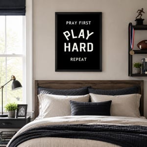 May include: A bedroom scene featuring a bed with a dark wooden headboard, layered with beige and black bedding, and decorative pillows. A framed black and white print on the wall reads "PRAY FIRST, PLAY HARD, REPEAT." A side table with a lamp and a small framed photo is visible.