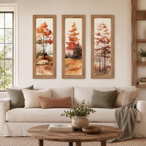 May include: Three framed watercolor paintings of autumn trees with orange and red foliage. The artwork is displayed above a white sofa with decorative pillows and a wooden coffee table. The room has a neutral color palette.