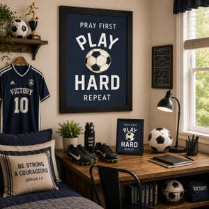 May include: A framed wall poster with a dark blue background featuring the white text PRAY FIRST PLAY HARD REPEAT and a soccer ball graphic. The room decor includes a navy soccer jersey, a desk with a smaller framed print, a soccer ball, and cleats.