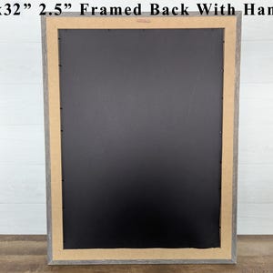May include: A framed artwork's back, measuring 26x32 inches with a 2.5-inch frame, is displayed. The frame has a gray finish, and the back is black. The artwork includes a hanger for easy wall mounting. The frame is on a wooden surface.