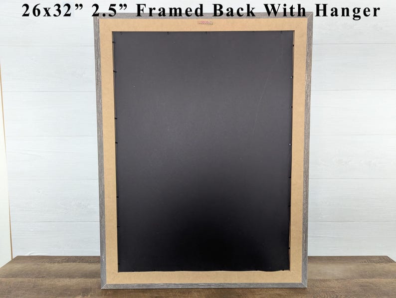 May include: A framed artwork's back, measuring 26x32 inches with a 2.5-inch frame, is displayed. The frame has a gray finish, and the back is black. The artwork includes a hanger for easy wall mounting. The frame is on a wooden surface.