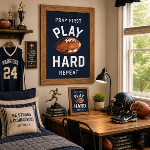 May include: A framed wall art print featuring a dark blue background with the white text PRAY FIRST, PLAY HARD, REPEAT and a brown football illustration. The print is displayed in a wooden frame above a desk with sports-themed decor and accessories.