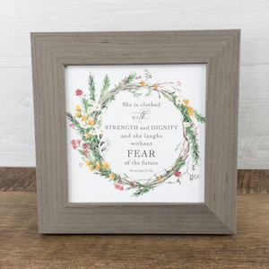 May include: A framed print with a floral wreath design and the text "She is clothed with STRENGTH and DIGNITY and she laughs without FEAR of the future Proverbs 31:25".