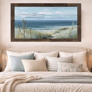 May include: A framed landscape art print depicting a serene beach scene with ocean waves, sand, and tall grass. The artwork has a wooden frame and includes the text "He leads me by still waters, He restores my soul, Psalm 23."