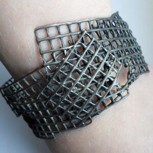 Könnte beinhalten: Ein silbernes Metallarmband mit geometrischem, durchbrochenem Design. Das Armband besteht aus miteinander verwobenen Metallstreifen, die ein gitterartiges Muster bilden.
