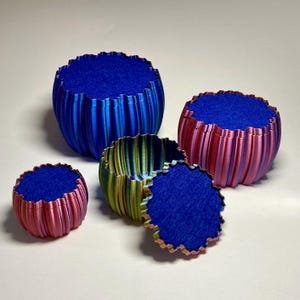 Könnte beinhalten: Ein Set aus vier runden, gerippten Behältern mit Farbverlauf. Ein großer blauer Behälter, ein mittlerer in Pink und Lila, ein kleiner in Grün und Gelb sowie ein passender Deckel. Jedes Teil ist mit einem leuchtend blauen Filz ausgekleidet.