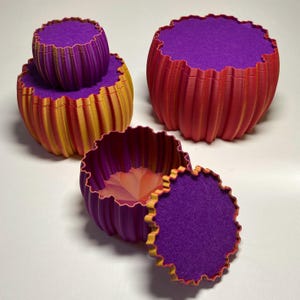 May include: Three round, ribbed containers with scalloped edges and purple felt-lined lids. One container is stacked on another, while the third sits open with its lid placed beside it. The exterior features a gradient of red, orange, and yellow.