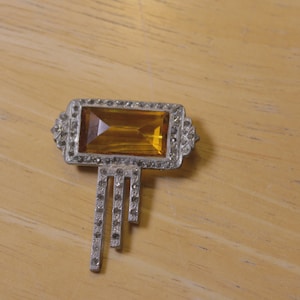 May include: A rectangular silver-tone brooch featuring a large, faceted amber-colored central stone. The frame is bordered by small, clear pavé-set crystals, with three vertical, stepped-length metal drops extending from the bottom, each accented with clear crystals.