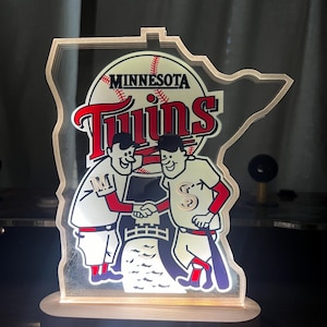 Puede incluir: Un logotipo iluminado del equipo de b&eacute;isbol Minnesota Twins con la forma del estado de Minnesota. El logotipo presenta dos jugadores de b&eacute;isbol de dibujos animados d&aacute;ndose la mano frente a una pelota de b&eacute;isbol y las palabras "Minnesota Twins".
