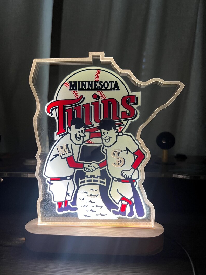 Puede incluir: Un logotipo iluminado del equipo de b&eacute;isbol Minnesota Twins con la forma del estado de Minnesota. El logotipo presenta dos jugadores de b&eacute;isbol de dibujos animados d&aacute;ndose la mano frente a una pelota de b&eacute;isbol y las palabras "Minnesota Twins".