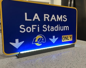 Könnte beinhalten: Ein blaues und gelbes Schild mit dem Text "LA RAMS SoFi Stadium" und einem nach unten zeigenden Pfeil. Das Schild hat eine schwarze Basis mit einem blauen Licht darunter.
