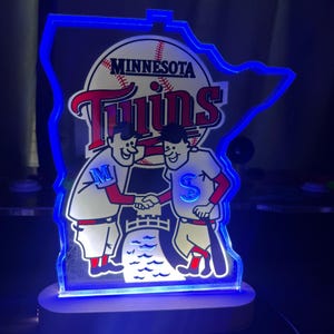 Puede incluir: Un letrero iluminado azul y blanco con forma del estado de Minnesota con las palabras "Minnesota Twins" y un gr&aacute;fico de b&eacute;isbol. Dos jugadores de b&eacute;isbol de dibujos animados se dan la mano frente a un bate de b&eacute;isbol.