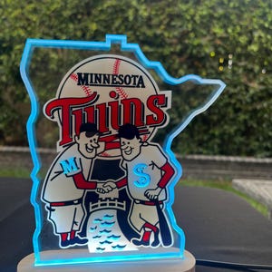 Puede incluir: Un letrero luminoso azul y blanco con forma del estado de Minnesota con las palabras "Minnesota Twins" y un gr&aacute;fico de b&eacute;isbol. Dos jugadores de b&eacute;isbol de dibujos animados se dan la mano frente a un bate de b&eacute;isbol y una pelota de b&eacute;isbol.