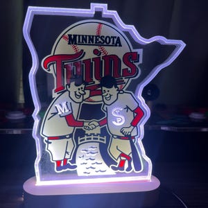 Puede incluir: Un logotipo blanco iluminado de los Minnesota Twins con la forma del estado de Minnesota. El logotipo presenta dos jugadores de b&eacute;isbol de dibujos animados d&aacute;ndose la mano frente a una pelota de b&eacute;isbol con la palabra "Minnesota" y la palabra "Twins" sobre la pelota.