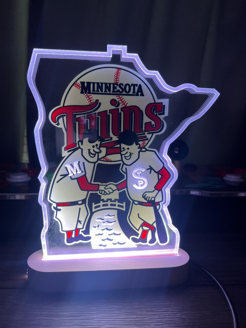 Puede incluir: Un logotipo blanco iluminado de los Minnesota Twins con la forma del estado de Minnesota. El logotipo presenta dos jugadores de b&eacute;isbol de dibujos animados d&aacute;ndose la mano frente a una pelota de b&eacute;isbol con la palabra "Minnesota" y la palabra "Twins" sobre la pelota.