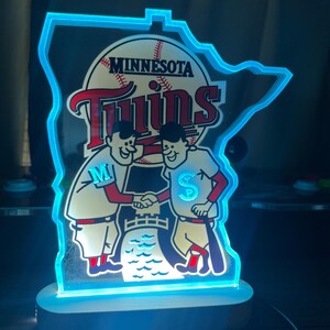 Puede incluir: Un letrero luminoso azul y blanco con forma del estado de Minnesota con el logotipo de los Minnesota Twins. El logotipo presenta una pelota de b&eacute;isbol, la palabra "Twins" y dos figuras de dibujos animados d&aacute;ndose la mano. Las figuras llevan uniformes de b&eacute;isbol con las letras "M" y "S" en el pecho.