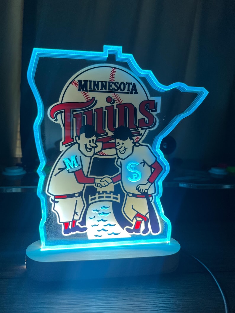 Puede incluir: Un letrero luminoso azul y blanco con forma del estado de Minnesota con el logotipo de los Minnesota Twins. El logotipo presenta una pelota de b&eacute;isbol, la palabra "Twins" y dos figuras de dibujos animados d&aacute;ndose la mano. Las figuras llevan uniformes de b&eacute;isbol con las letras "M" y "S" en el pecho.