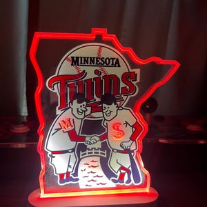 Puede incluir: Un logotipo rojo y blanco de los Minnesota Twins con dos jugadores de b&eacute;isbol de dibujos animados d&aacute;ndose la mano frente a una pelota de b&eacute;isbol. El logotipo est&aacute; sobre una base de acr&iacute;lico transparente con un contorno rojo. La base est&aacute; iluminada con luces LED rojas.