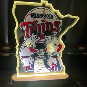 Puede incluir: Un logotipo iluminado de los Minnesota Twins con la forma del estado de Minnesota. El logotipo presenta a dos jugadores de b&eacute;isbol de dibujos animados d&aacute;ndose la mano frente a una pelota de b&eacute;isbol con la palabra "Twins" y "Minnesota" escrita en ella.
