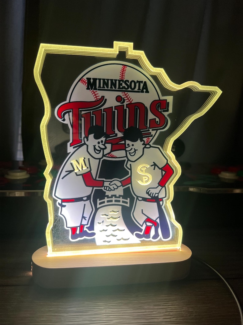 Puede incluir: Un logotipo iluminado de los Minnesota Twins con la forma del estado de Minnesota. El logotipo presenta a dos jugadores de b&eacute;isbol de dibujos animados d&aacute;ndose la mano frente a una pelota de b&eacute;isbol con la palabra "Twins" y "Minnesota" escrita en ella.