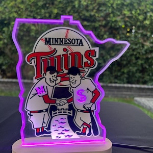 Puede incluir: Una l&aacute;mpara iluminada de los Minnesota Twins de color morado y blanco con forma del estado de Minnesota. La l&aacute;mpara presenta dos jugadores de b&eacute;isbol de dibujos animados d&aacute;ndose la mano con el logotipo del equipo y las palabras "Minnesota Twins" encima.