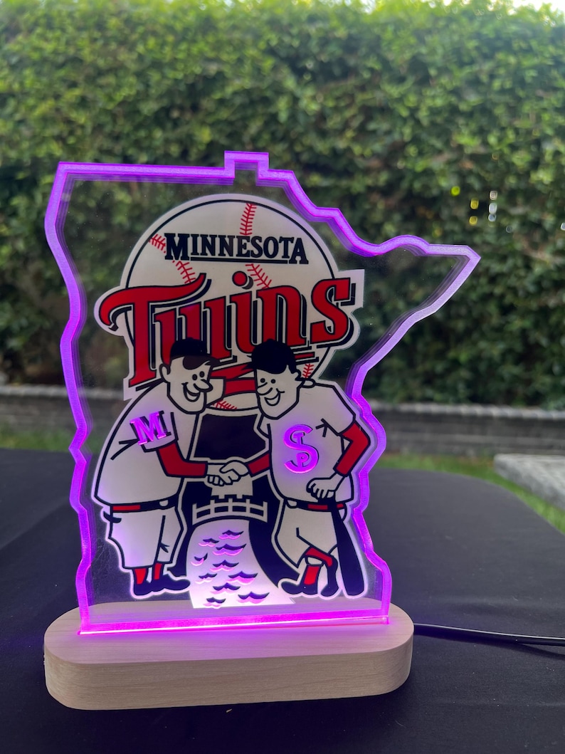 Puede incluir: Una l&aacute;mpara iluminada de los Minnesota Twins de color morado y blanco con forma del estado de Minnesota. La l&aacute;mpara presenta dos jugadores de b&eacute;isbol de dibujos animados d&aacute;ndose la mano con el logotipo del equipo y las palabras "Minnesota Twins" encima.