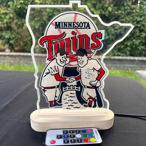 Puede incluir: Una luz nocturna de acr&iacute;lico transparente con el logotipo de los Minnesota Twins, con una base de madera y un control remoto. El logotipo presenta dos jugadores de b&eacute;isbol de dibujos animados d&aacute;ndose la mano frente a una pelota de b&eacute;isbol y las palabras "Minnesota Twins".