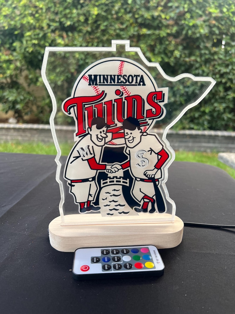 Puede incluir: Una luz nocturna de acr&iacute;lico transparente con el logotipo de los Minnesota Twins, con una base de madera y un control remoto. El logotipo presenta dos jugadores de b&eacute;isbol de dibujos animados d&aacute;ndose la mano frente a una pelota de b&eacute;isbol y las palabras "Minnesota Twins".
