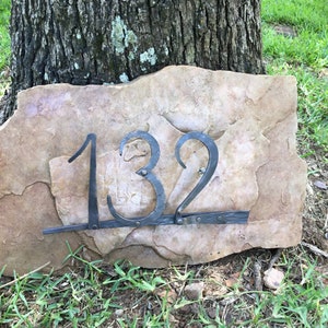 May include: A rustic address plaque featuring the number 132 in wrought iron against a natural stone backdrop. The numbers are dark gray and attached to a rectangular stone with a rough, textured surface. The plaque is set on grass.