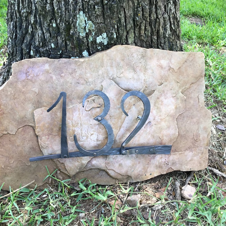 May include: A rustic address plaque featuring the number 132 in wrought iron against a natural stone backdrop. The numbers are dark gray and attached to a rectangular stone with a rough, textured surface. The plaque is set on grass.